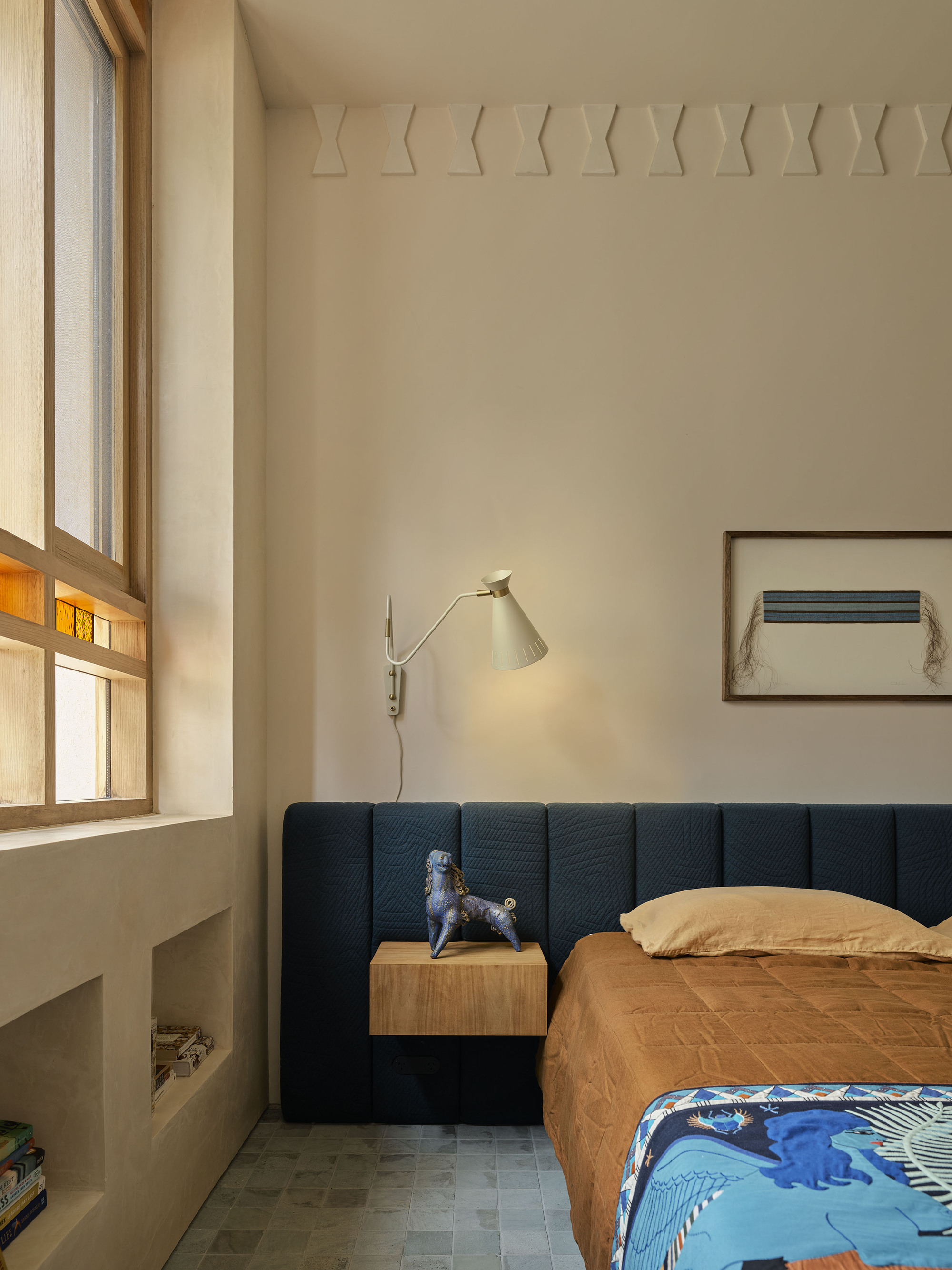 modern bedroom with hempcrete walls and modern wall molding, extended upholstered headboard with orange bedcover and colorful blanket, floating timber side table with statue, wall light, artwork above bed, and timber framed windows with stained glass detail and alcove niches built into wall
