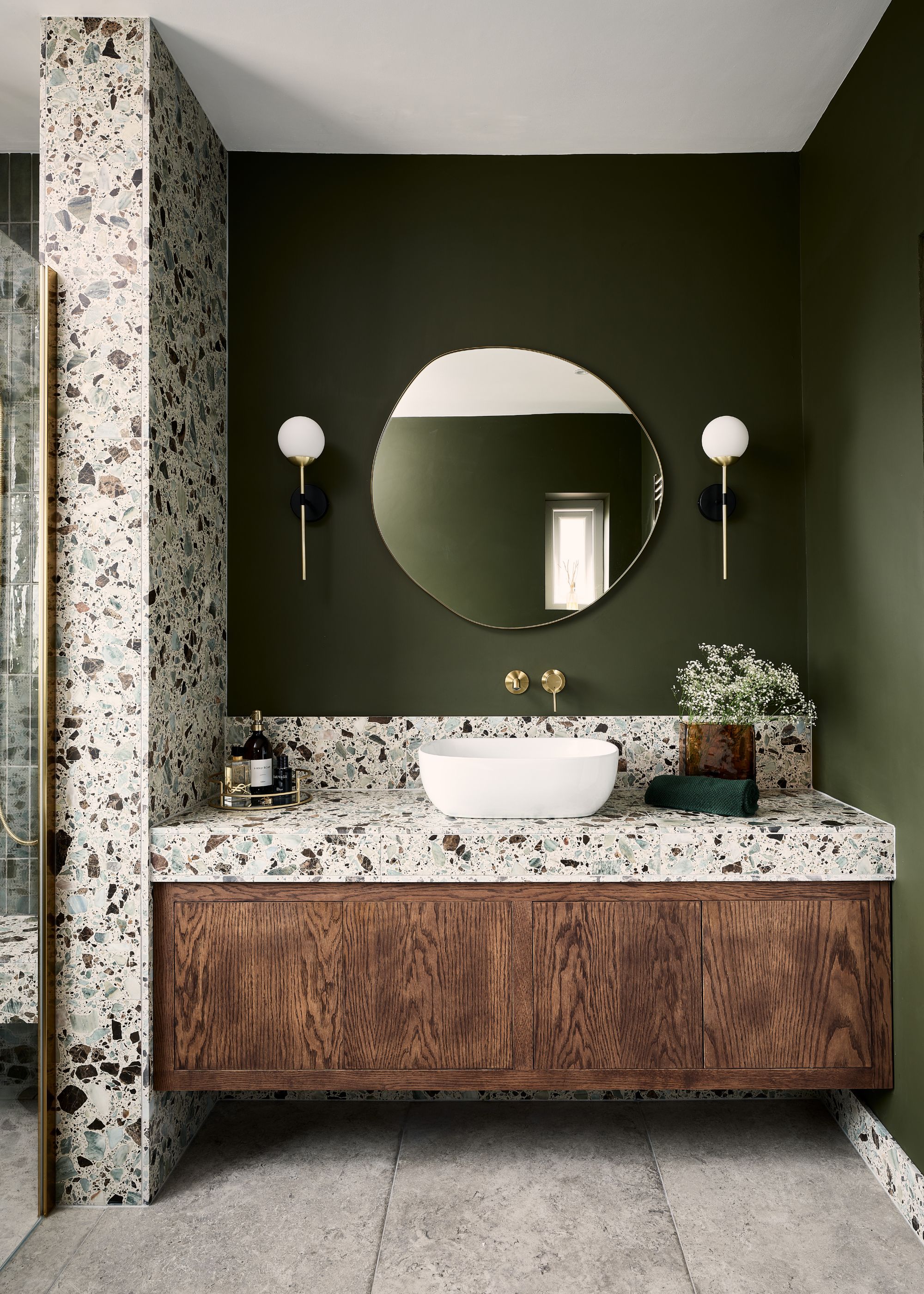 A bathroom with large terrazzo tiles used on the countertop and walls. Above the sink is a large curved mirror with lights on either side and on the vanity is a few bathroom essentials.