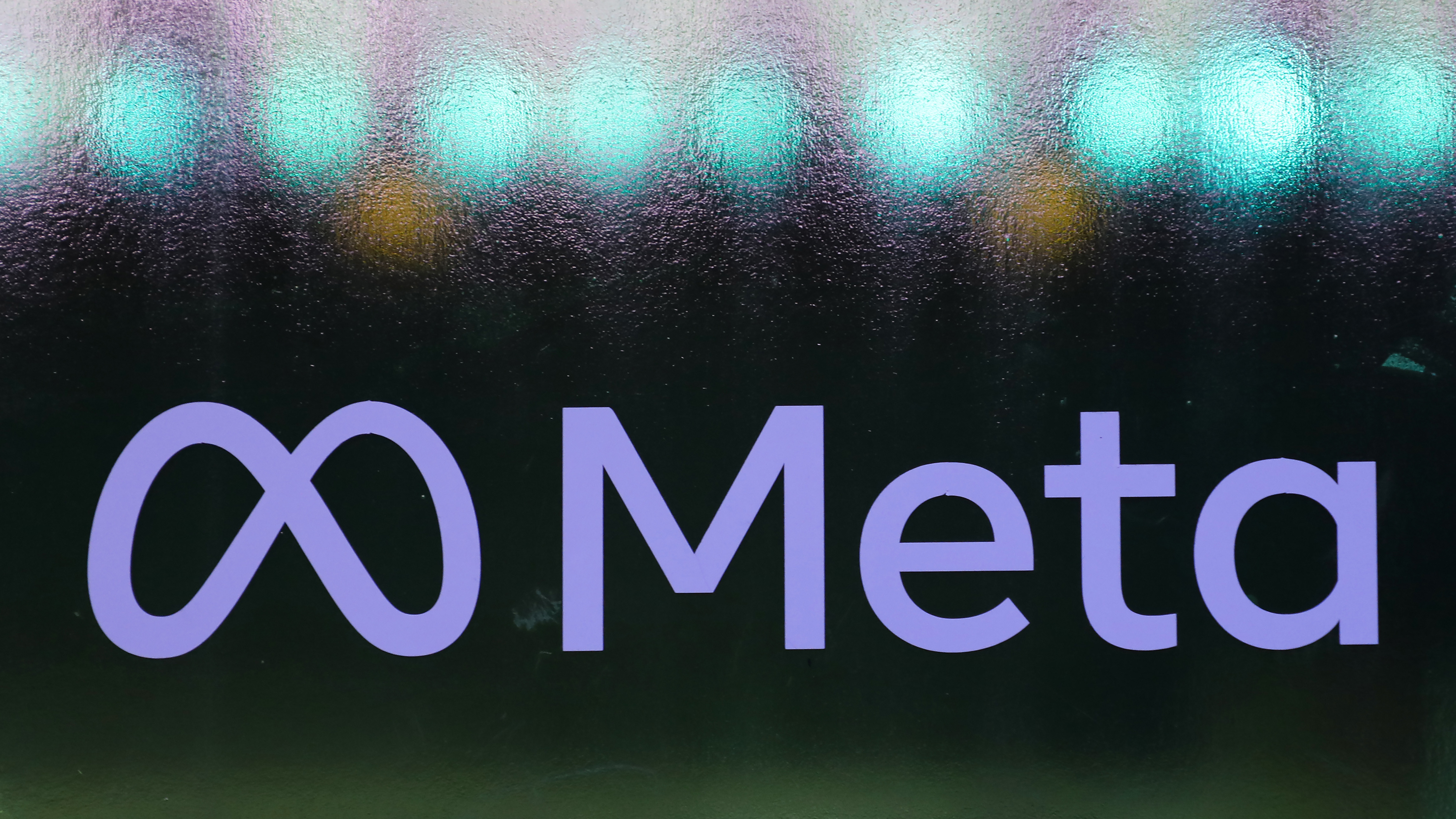 The Meta logo (a stylized 'M' next to the word 'Meta) written in blue on a frosted glass window, with blue lights visible behind the glass.