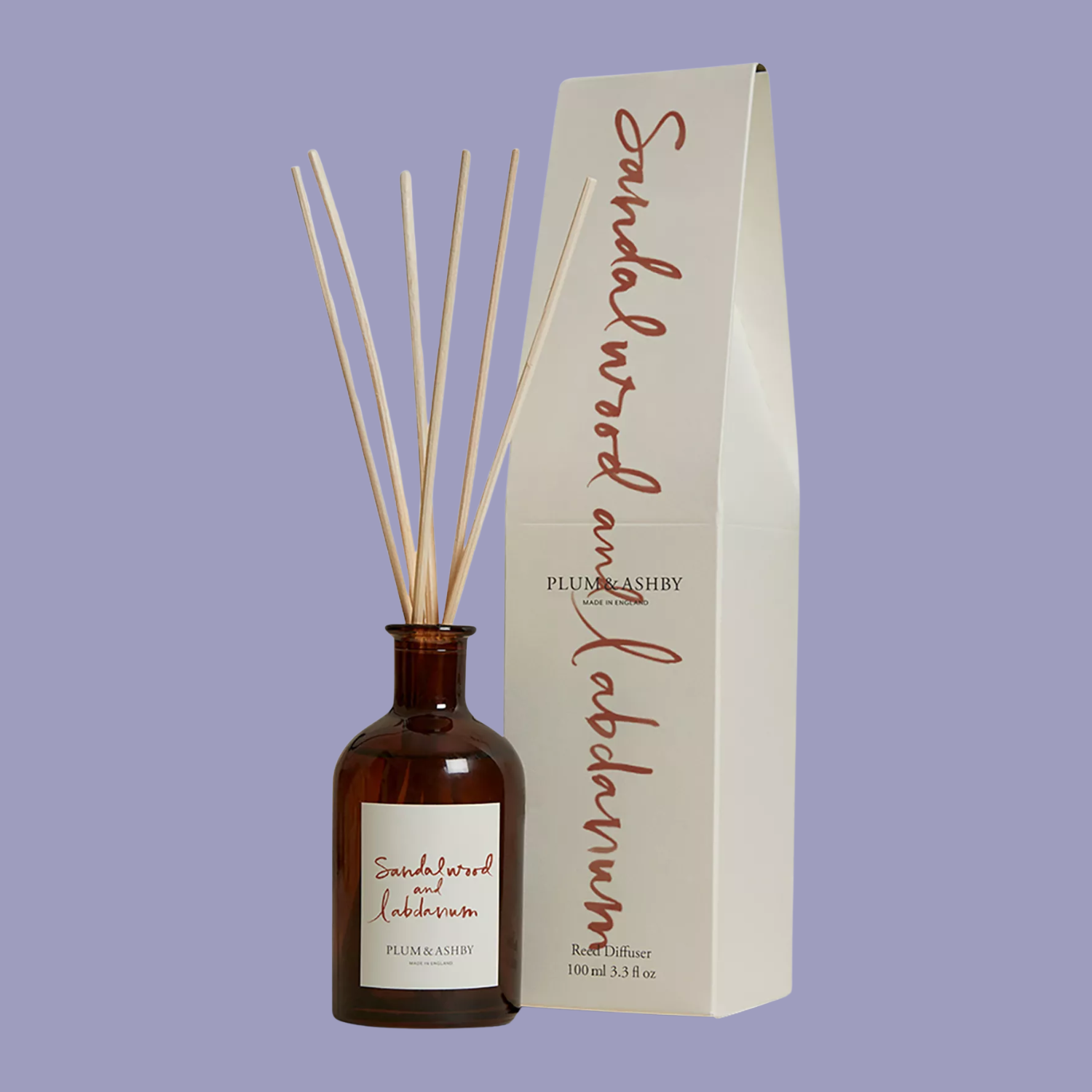 An amber glass reed diffuser and package against a purple backdrop