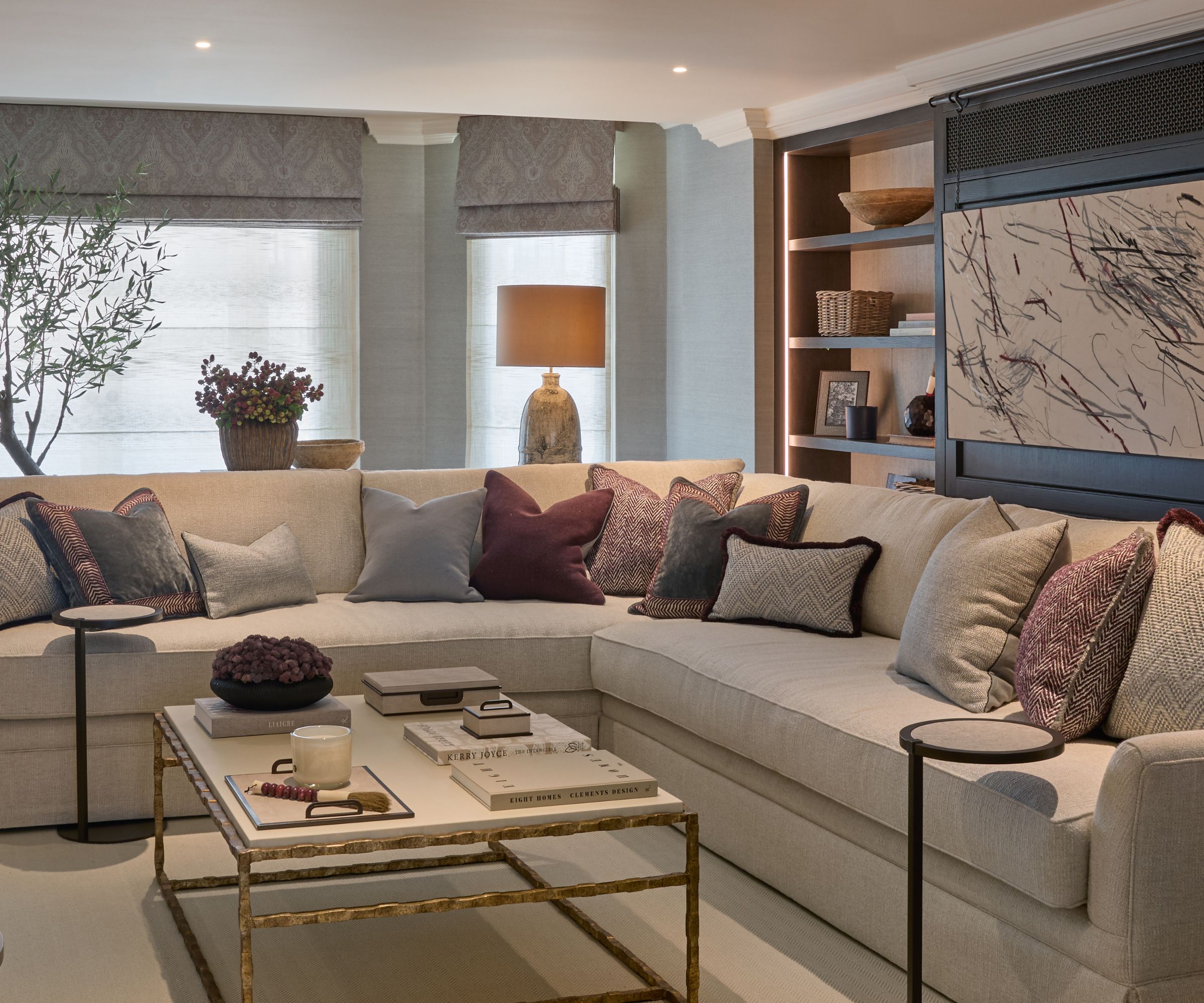 Corner of modern living room with large white corner sofa, shelving and artwork in background, cream carpet and square marble and metal coffee table in front of sofa, lamps and ornaments in back