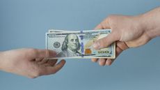 A man's hand holds out several hundred-dollar bills to the hand of a younger person. 