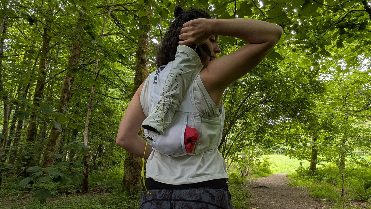 A woman trail runner in the forest pulling her poles out of a white Hoka hydration vest