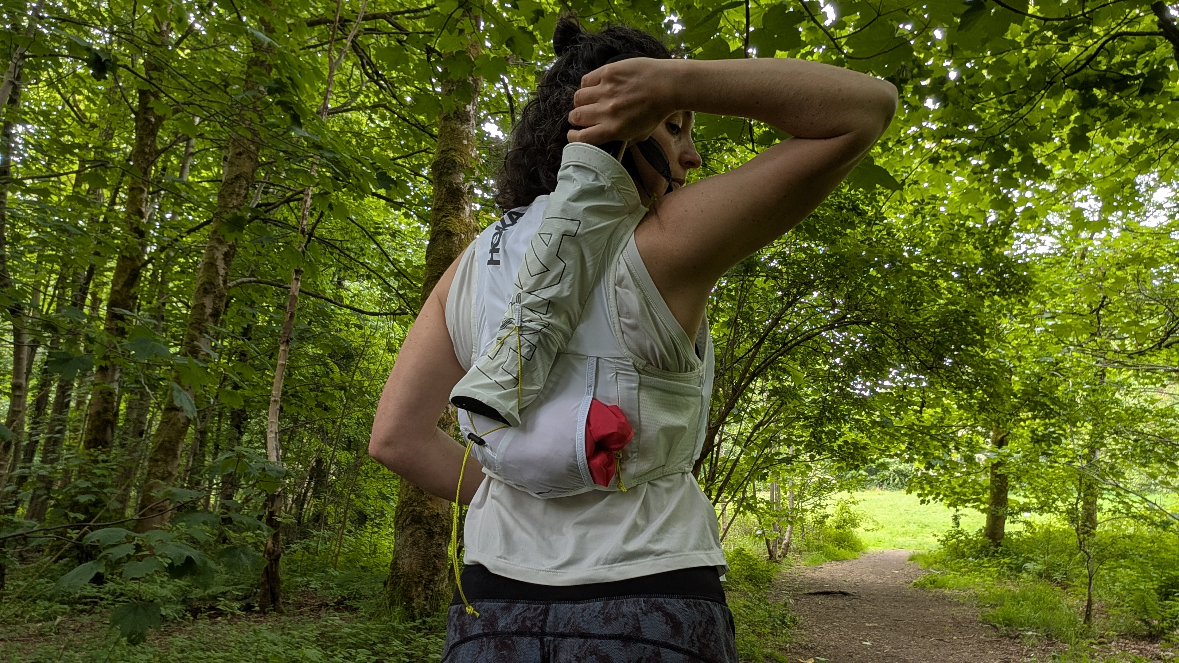 A woman trail runner in the forest pulling her poles out of a white Hoka hydration vest