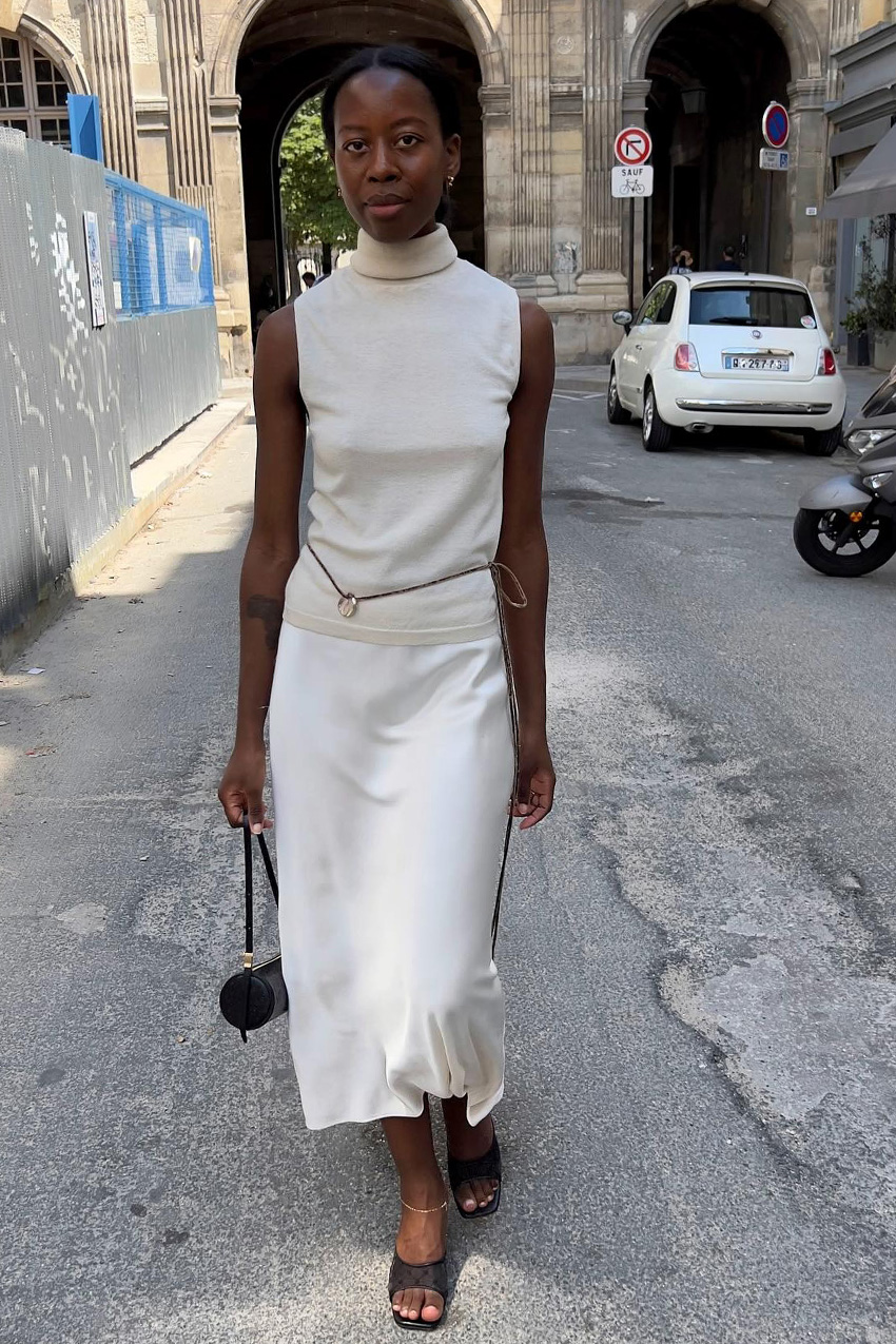How to wear a slip skirt is shown in a photo of a woman slip skirt outfit where she is walking down the street in Paris wearing gold earrings, a white turtleneck, a brown rope belt, a white slip skirt, a dainty gold anklet, black heeled mules, and a black barrel bag