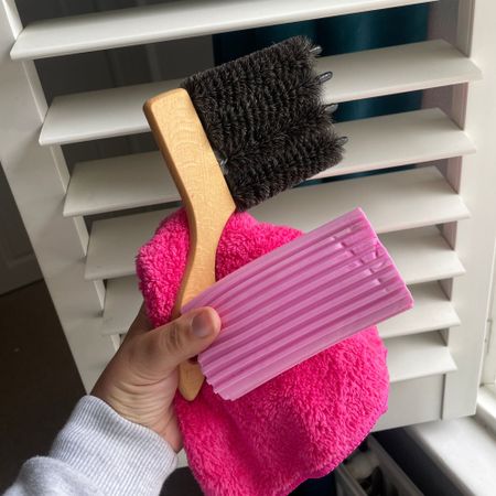 Hand holding a blind cleaning brush, a duster, and a Damp Duster in front of plantation shutters