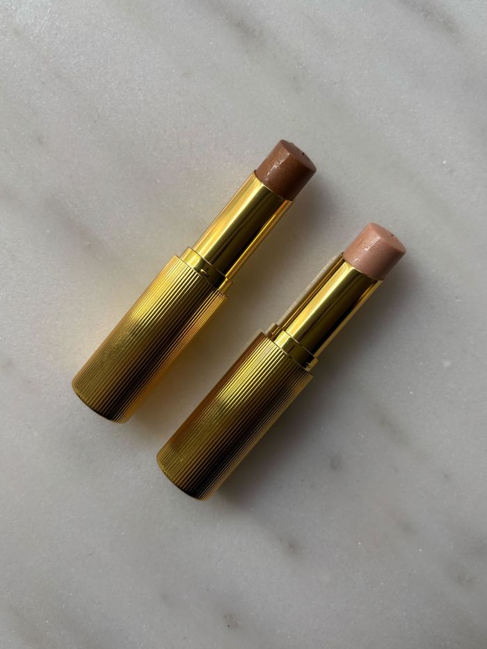 The Victoria Beckham Reflect Highlighter Stick in both shades on a marble table