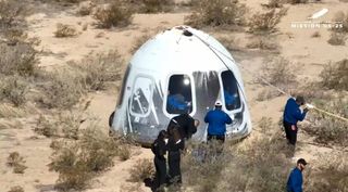 technicians in black jumpsuits walk around a white cone-shaped capsule on the desert floor