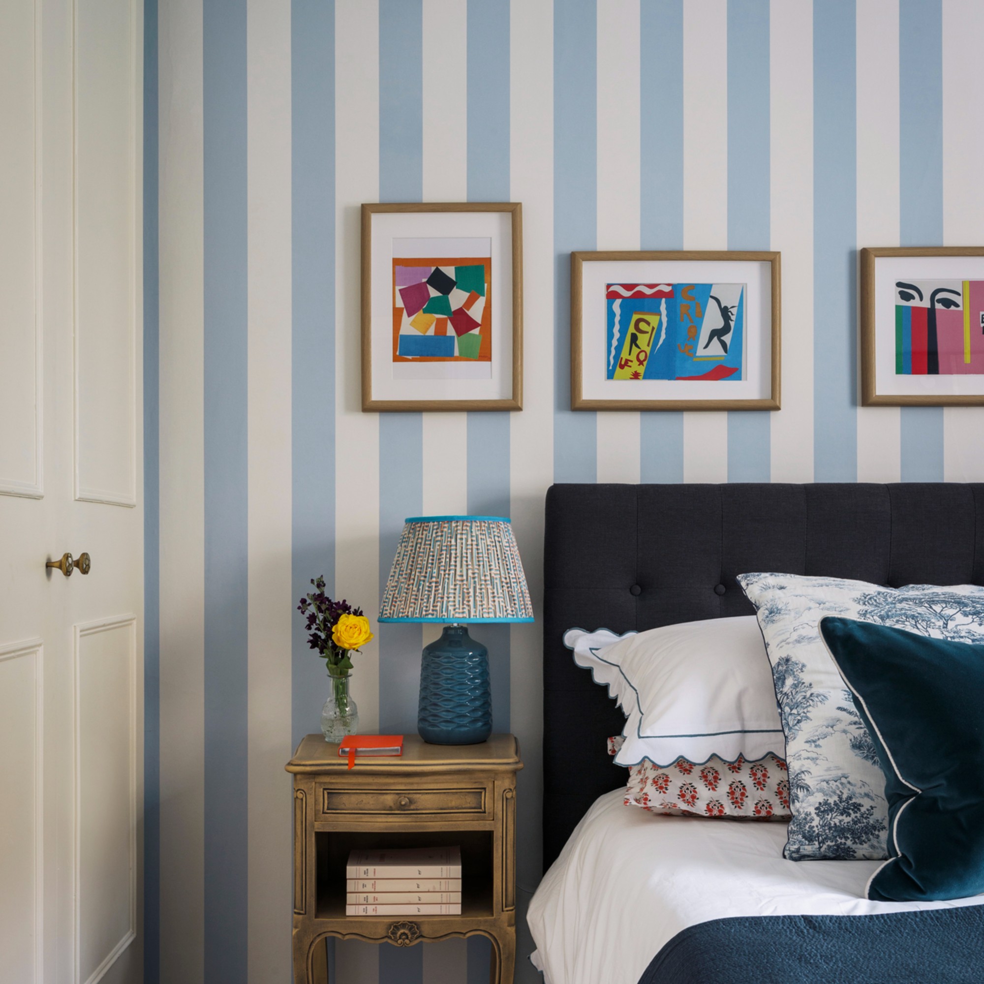 A bedroom with the wall behind the bed headboard covered in a soft blue wide striped wallpaper