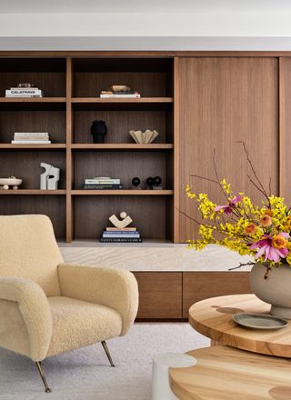 living room with boucle armchair, timber nesting coffee table with vase of flowers, and built-in cabinetry with open shelving styled with books and sculpture