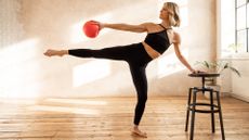 Tara Riley performs a Pilates move in a studio, standing on her left leg, with her right leg out to the side, and holding onto a stool for balance. Her right arm is clutching a Pilates ball and we see large windows and a bare wall behind her.