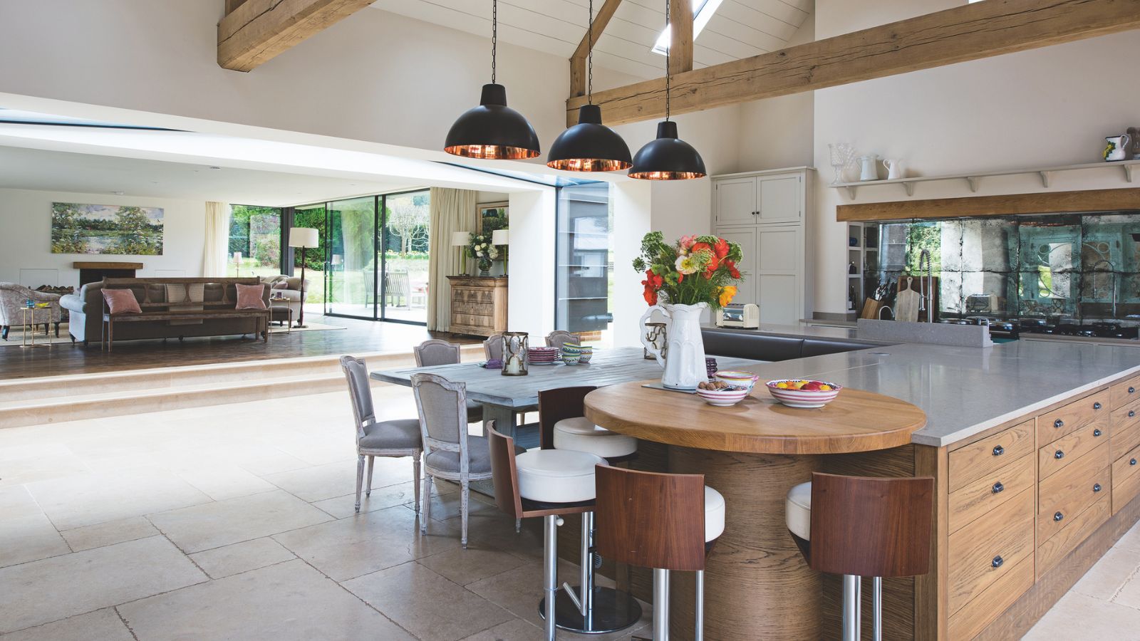 A spacious kitchen with tiled flooring and exposed wood beams. In the foreground is a large brown cabinet island with silver countertops, a circular wooden table, and a white dining table and chairs. On the island is a vase of flowers. In the background is a living space with a brown leather sofa, and large window doors out onto a green garden.