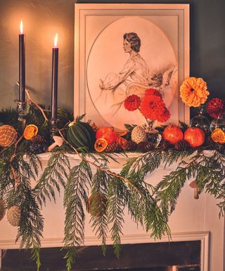 A mantlepiece with green leaf garland, small white and orange pumpkins, two lit tapered candles, and a framed illustration of a regency lady