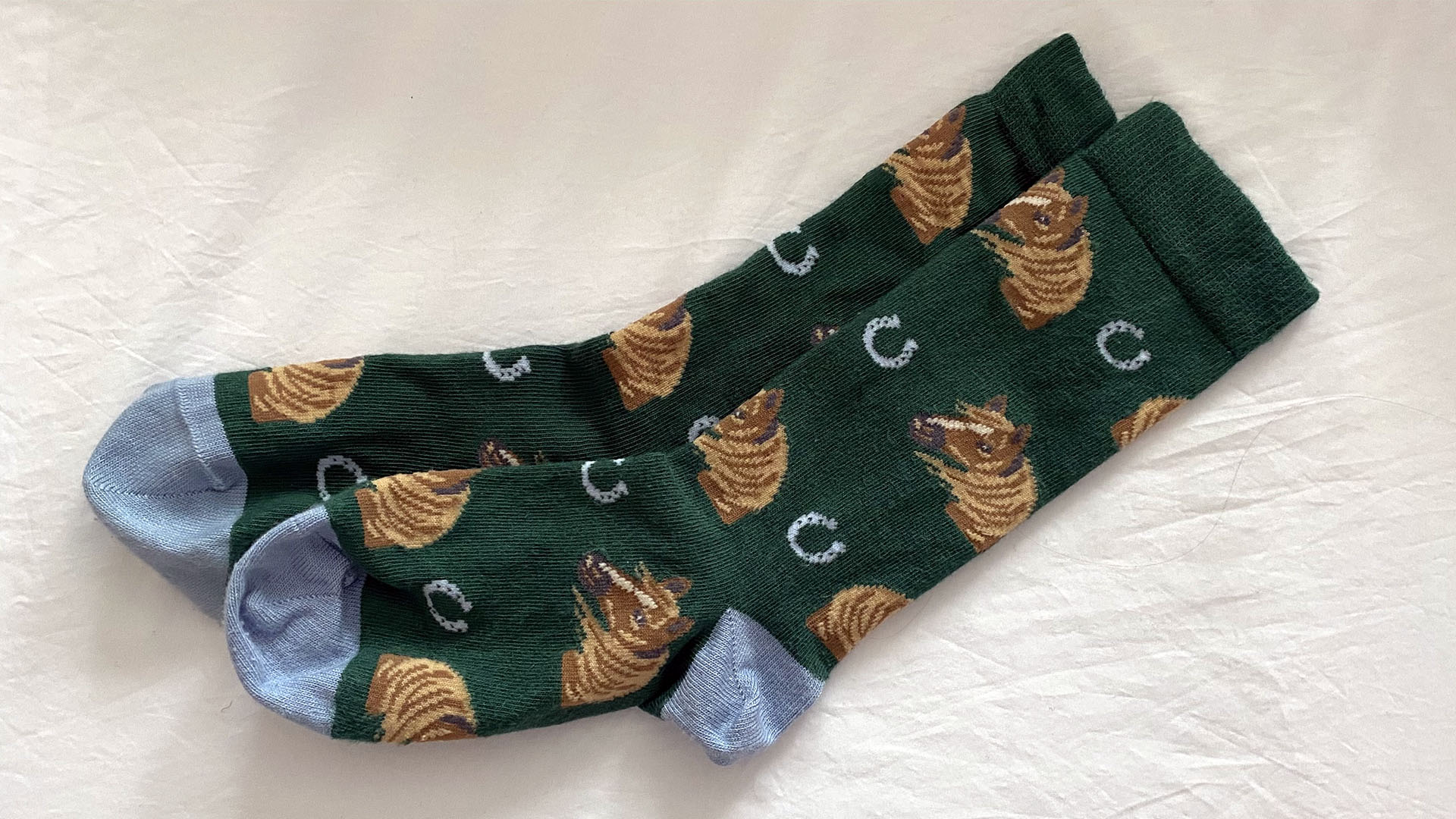 A pair of green socks patterned with horses and horse shoes, against white bed sheets