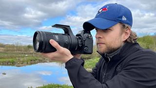 Mike Harris holding Nikon Z 70-200mm f/2.8 VR S II by a lake with vegetation and trees in the background 