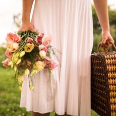 Woman wearing a white dress, holding a wicker picnic basket and a bouquet of roses