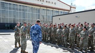 three men in camouflage uniforms address a crowd of people also in camouflage uniforms outside a hangar on a concrete tarmac