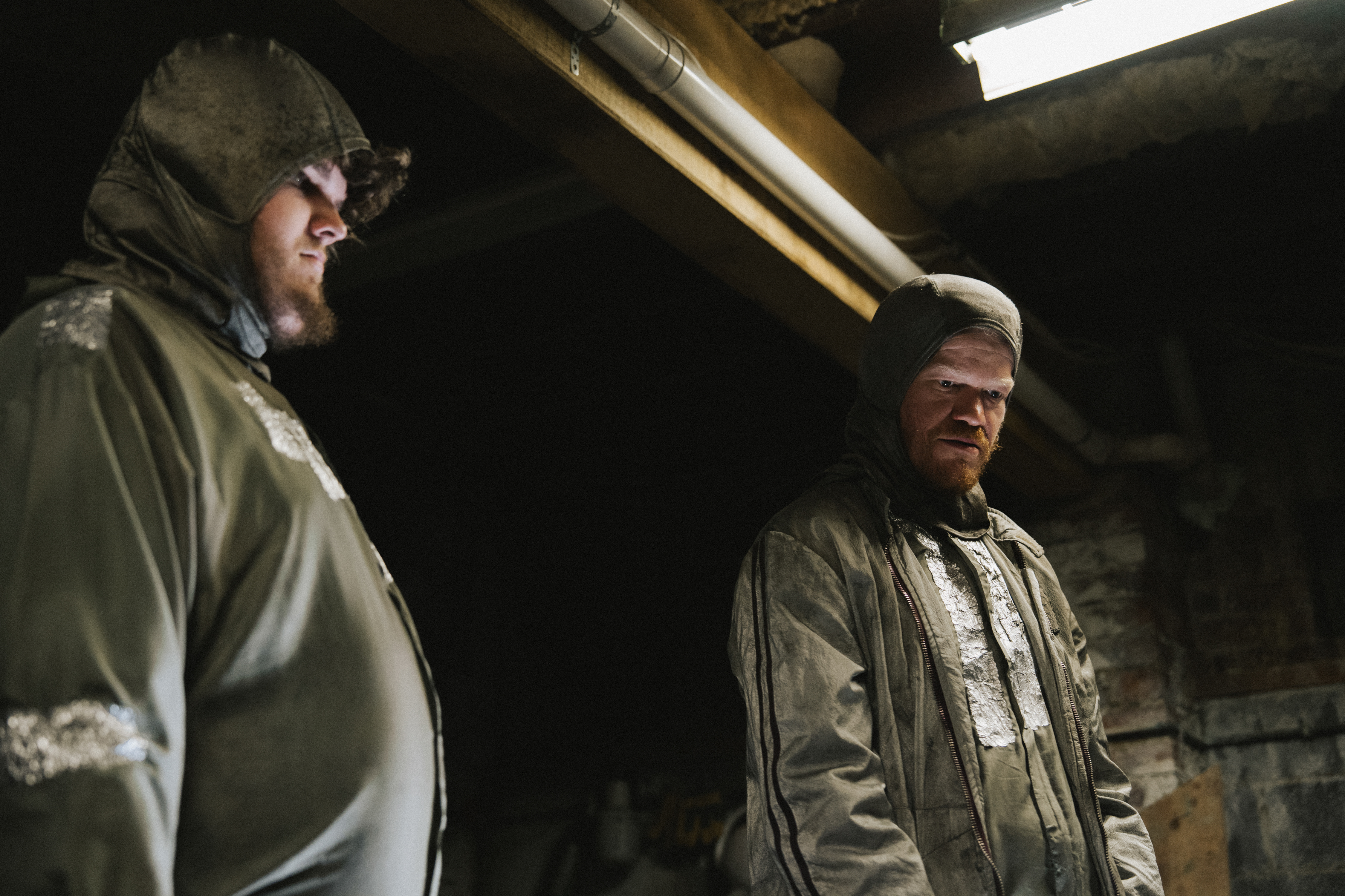 aidan delbis as don and jesse plemons as teddy wearing silver hazmat suits in a basement in bugonia