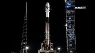 a rocket stands upright on launch pad at night with a large white payload atop it
