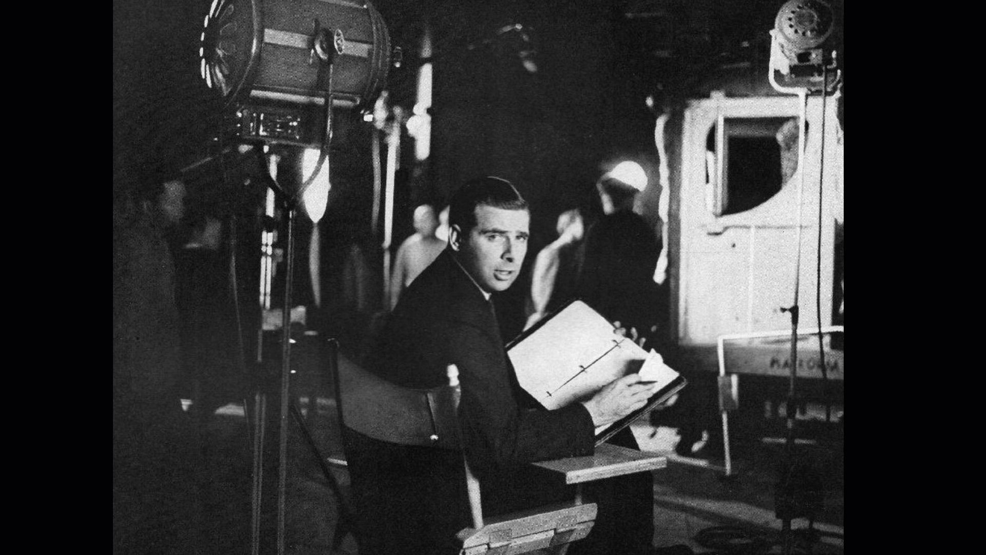 A black and white photo shows a man sitting in the director's chair with his back toward the camera next to a light with his face turned to the camera looking at a piece of paper
