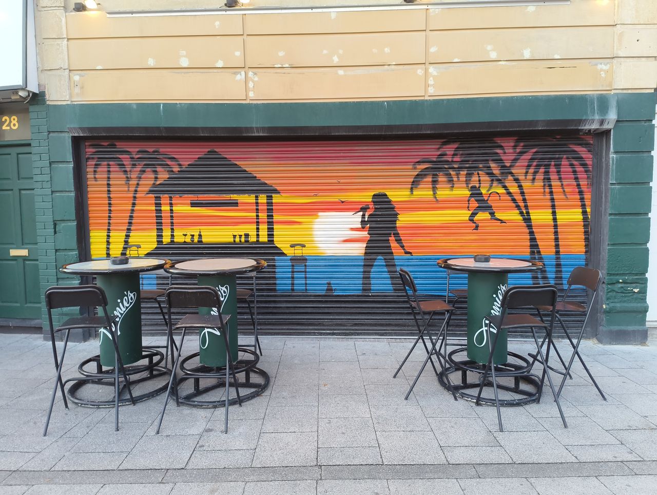 Two small outdoor caf&eacute; tables with matching chairs are set up in front of a closed storefront with a vibrant mural depicting a tropical sunset scene, a silhouetted singer, and palm trees.