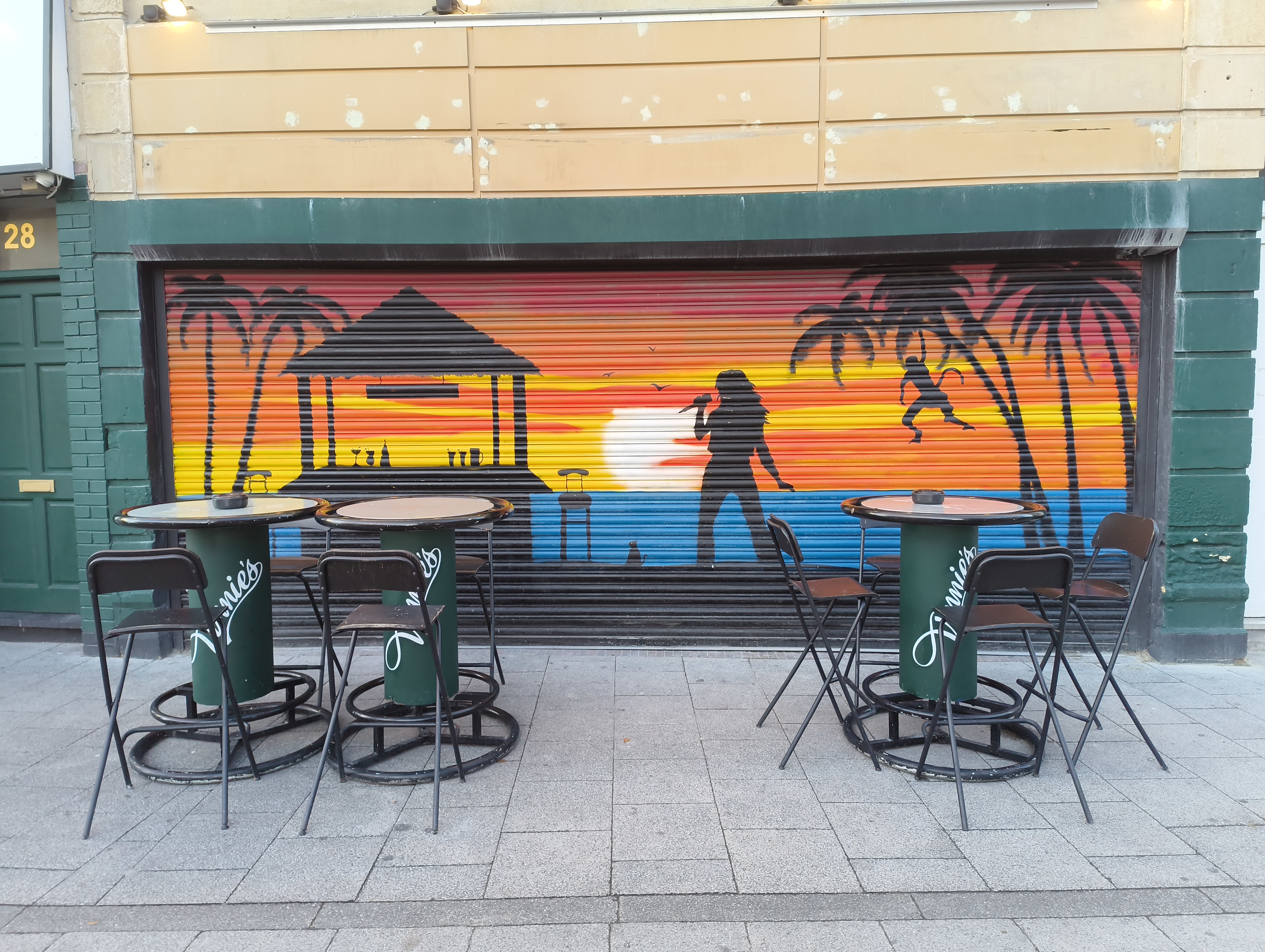 Two small outdoor café tables with matching chairs are set up in front of a closed storefront with a vibrant mural depicting a tropical sunset scene, a silhouetted singer, and palm trees.