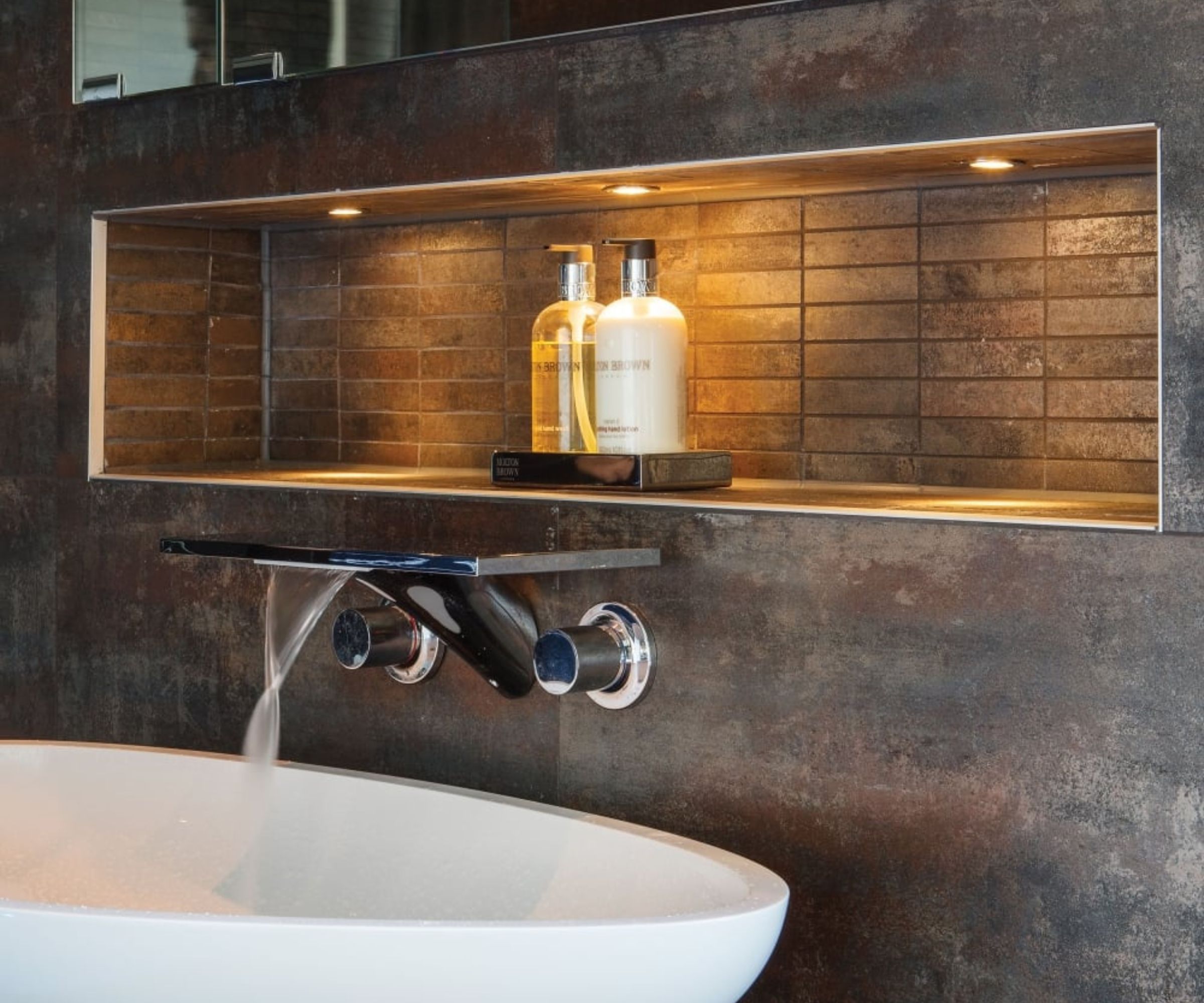 A close-up of a modern bathroom sink area. Above the sleek white basin is a wall-mounted waterfall-style tap with chrome handles, pouring a gentle stream of water. Behind the sink, a recessed shelf is built into the wall, lined with textured brown tiles