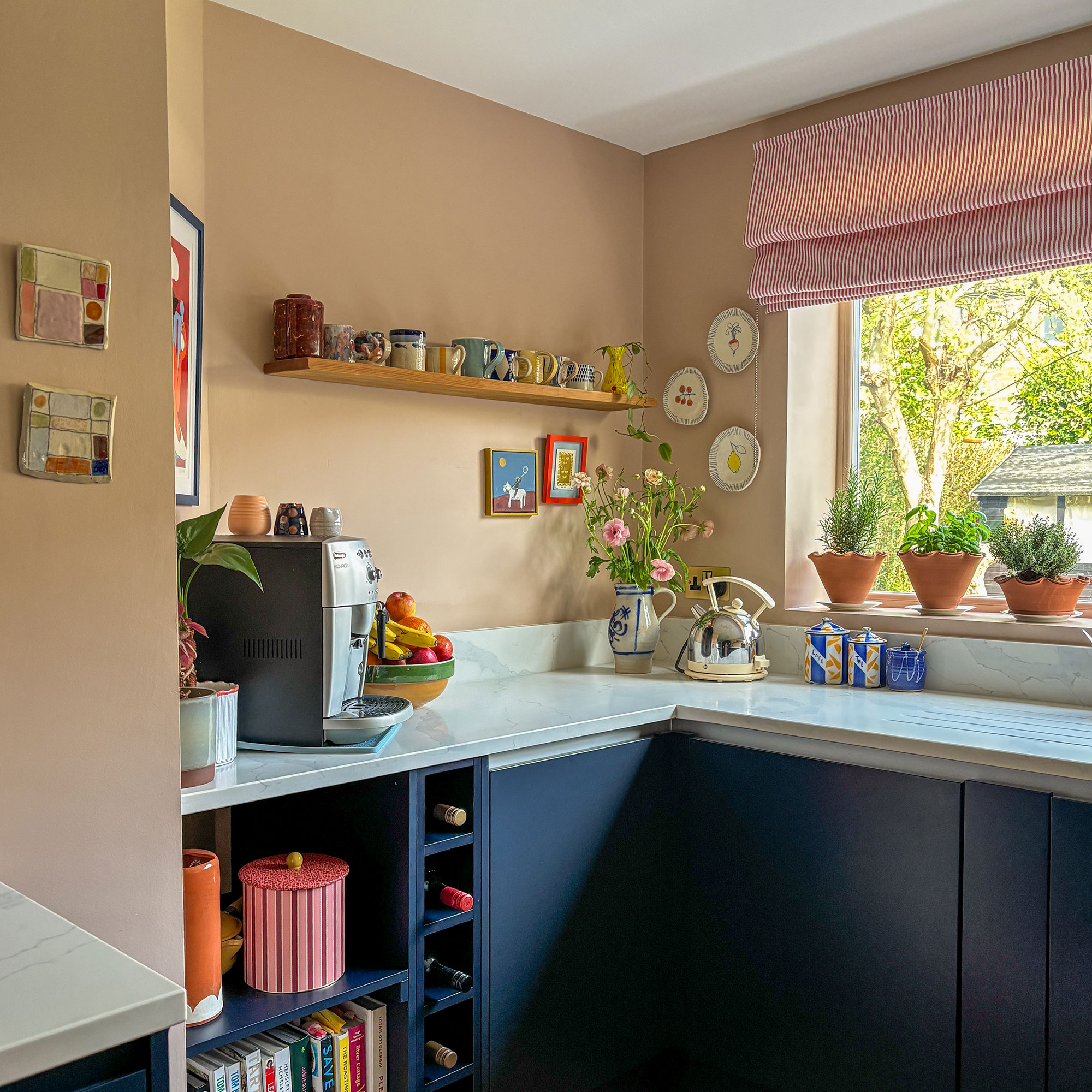 Peach kitchen walls above stone worktop and blue cabinetry, with window, coffee machine, kettle, potted herbs and floating shelf
