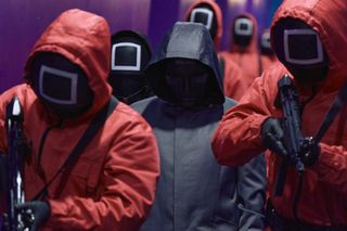 A man in a black hooded coat with his face obscured (Lee Byung-hun as Front Man) stands surrounded by red-suited square-faced guards holding guns, in 'Squid Game' season 2.