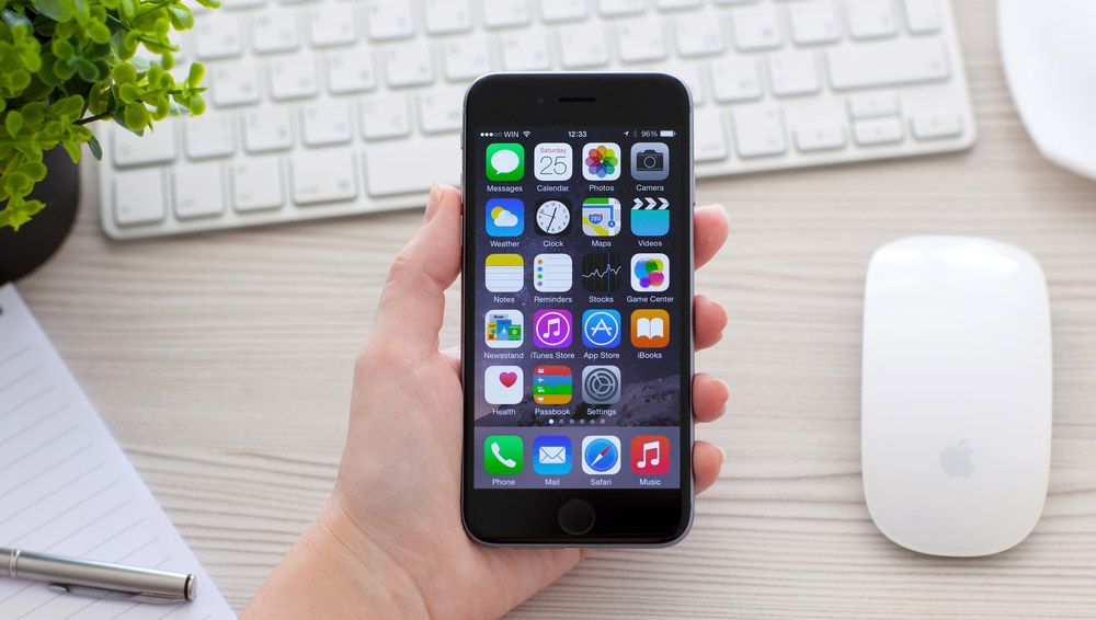 A hand holding a brand-new iPhone 6 over an Apple Magic Mouse and iMac keyboard.