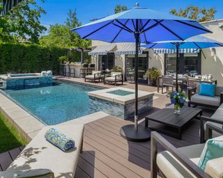 A modern swimming pool surrounded by decking, a living room area, dining zone and a garden bar, with blue umbrellas.