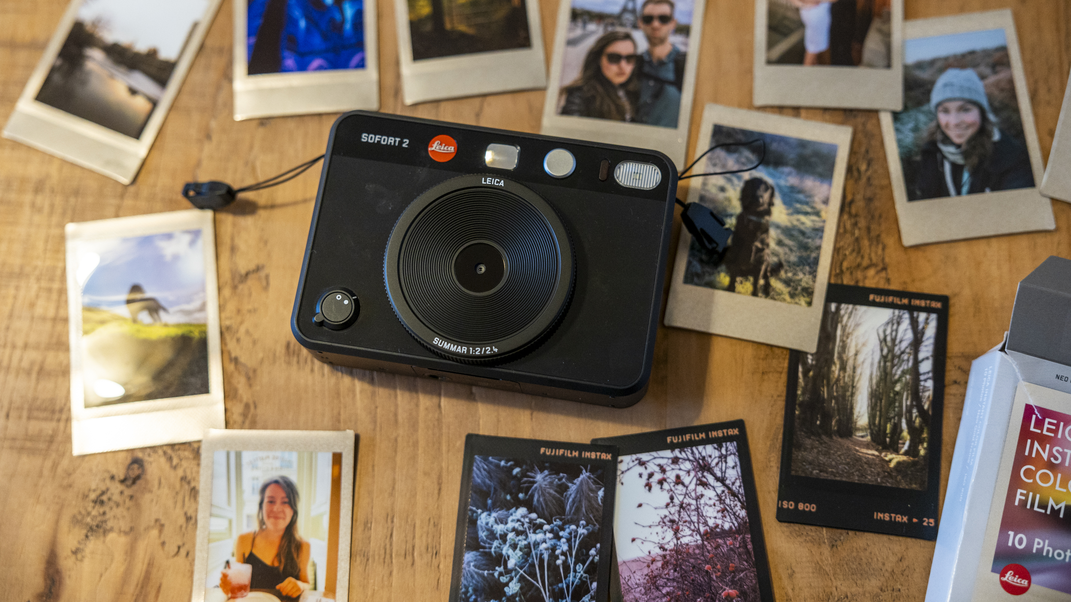 The Leica Sofort 2 on a wooden table surrounded by Instax prints