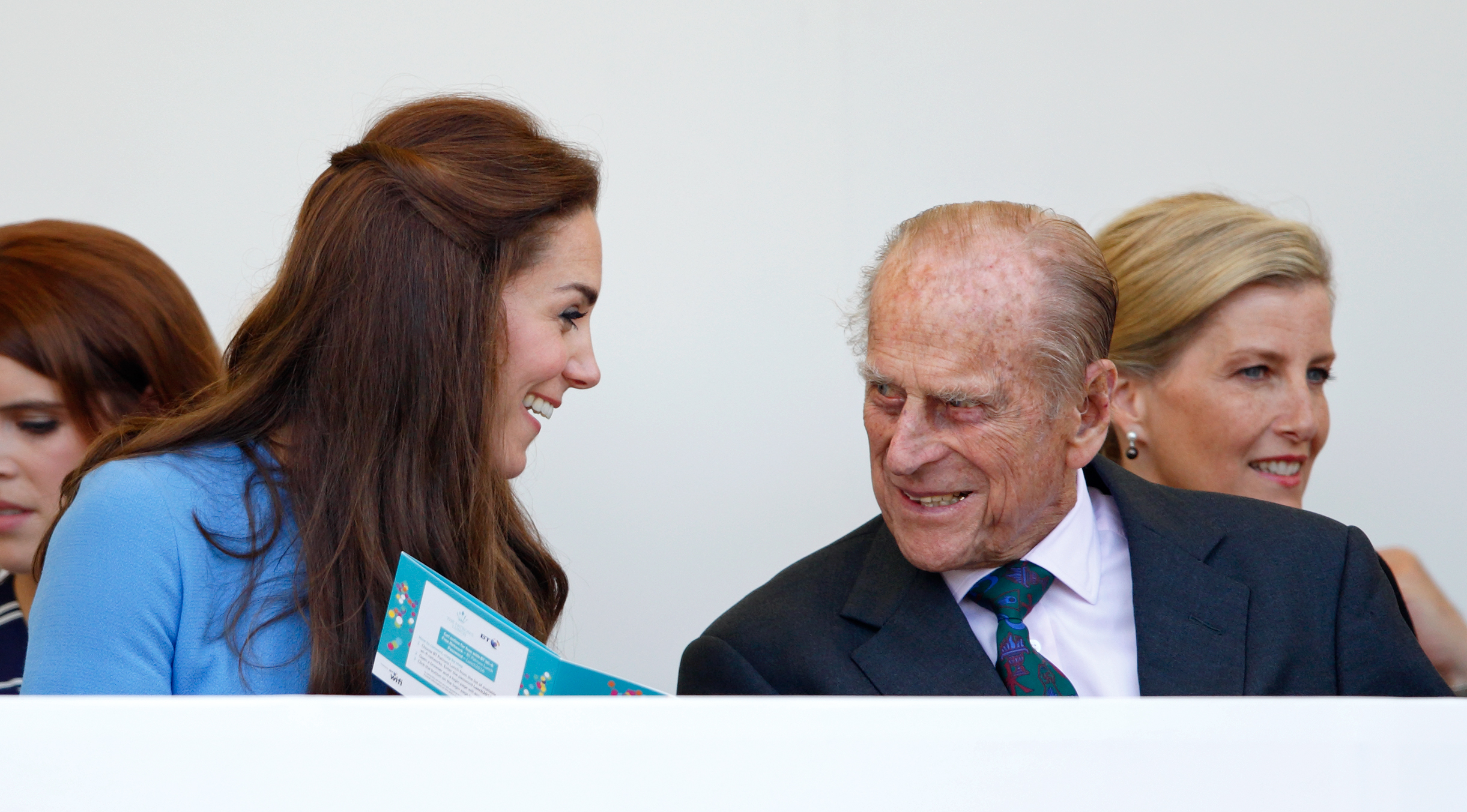 Princess Kate wearing a blue dress and Prince Philip in a suit and tie sitting next to each other, talking and laughing