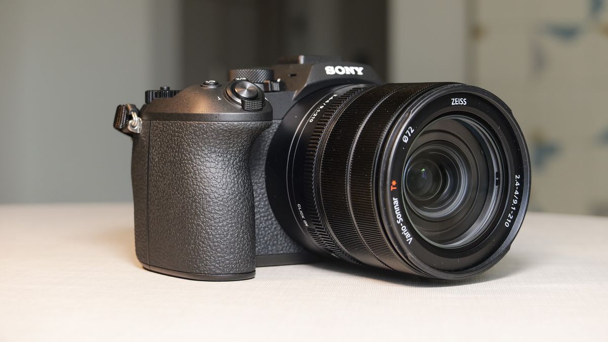 The Sony RX10 V bridge camera on a white table with a patterned wallpaper background, from the side of its grip