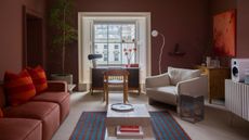 red living room with red sofa, red and orange cushion, blue and red striped rug, desk under a window, white armchair in front of a bar with artwork above and console table with speaker