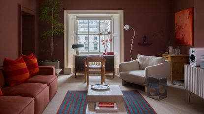 red living room with red sofa, red and orange cushion, blue and red striped rug, desk under a window, white armchair in front of a bar with artwork above and console table with speaker