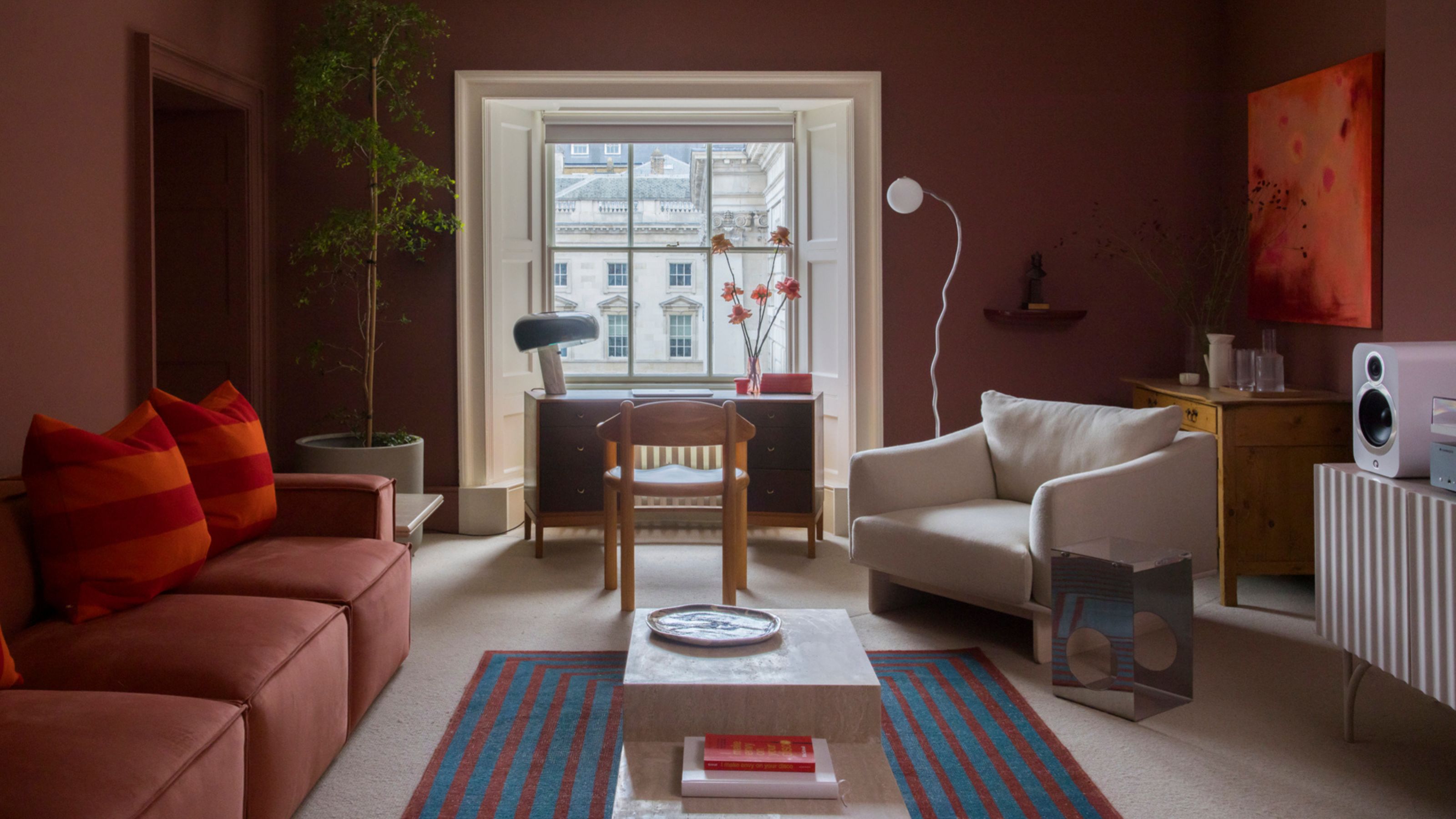 red living room with red sofa, red and orange cushion, blue and red striped rug, desk under a window, white armchair in front of a bar with artwork above and console table with speaker