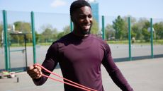 A smiling man in a long-sleeved top stands outside on a concrete sports pitch, stretching a resistance band between his hands. 