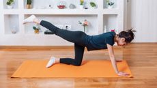 A woman in a t-shirt, leggings and socks works out on a yoga mat in a living room. Both her hands are on the floor and she is looking down towards the mat. Her left knee and shin are on the ground, while her right leg is extended behind her, parallel with the ground. Behind her are some shelves filled with decorative items; mostly plants, but some ceramics too. 