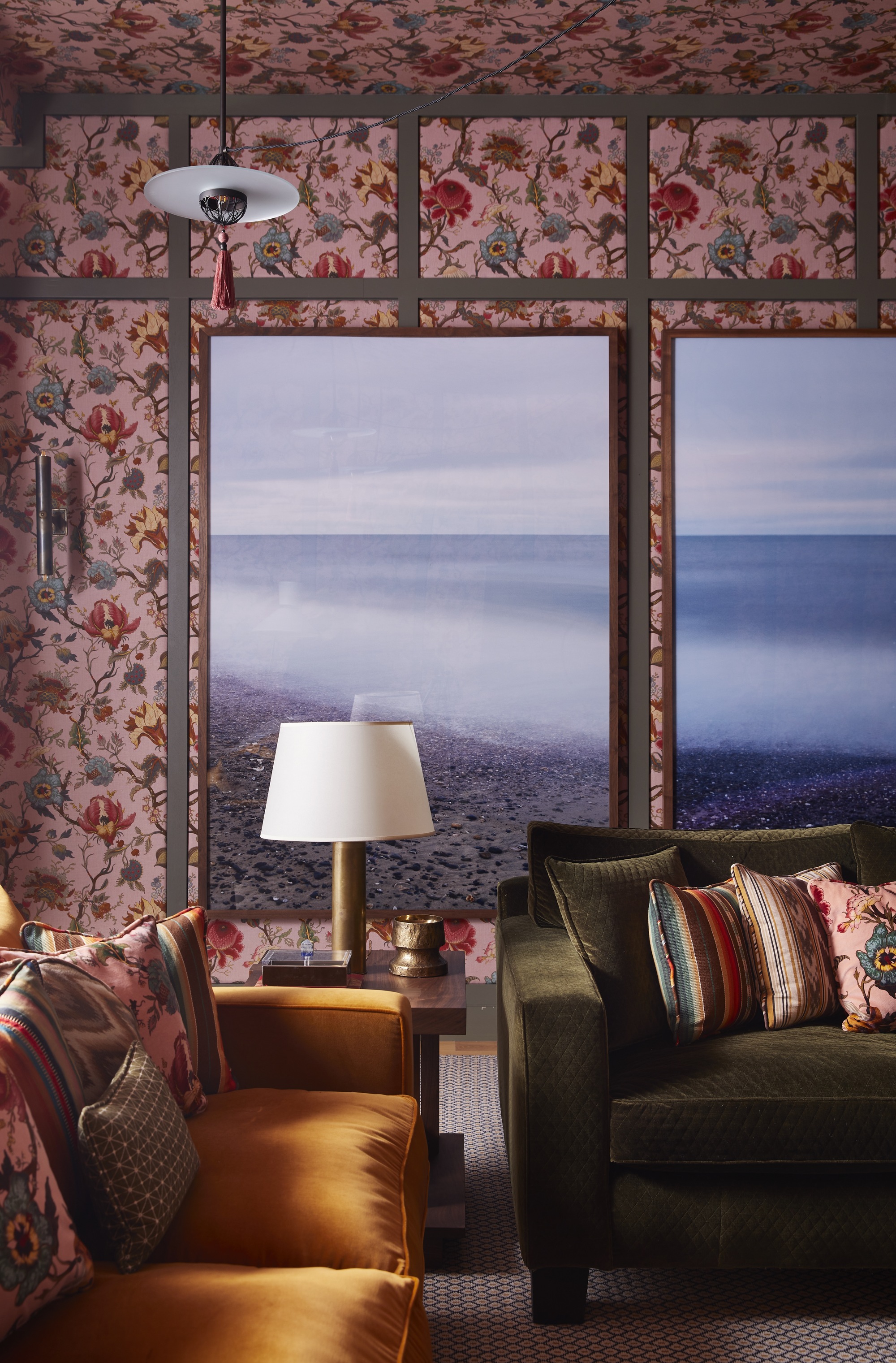living room with pink floral wallpaper, large artworks of the beach, corner of green velvet sofa and mustard yellow sofa, both decorated with colorful cushions, and side table in between with a lamp