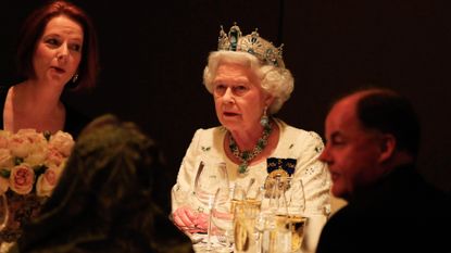 Queen Elizabeth wearing a tiara, white dress and diamond necklace sitting at a table in the dark