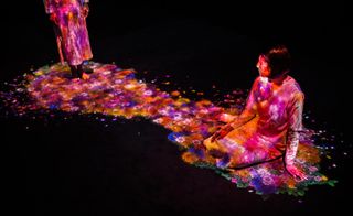 Art installation of TeamLab in the Pace Gallery. The room is dark. A woman is sitting on the floor while the. Digital art representing bloomed flowers covers the woman.