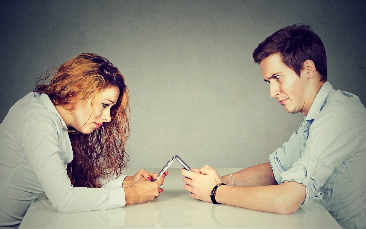 A man and woman sit across table from each other, each looking at their smartphone.