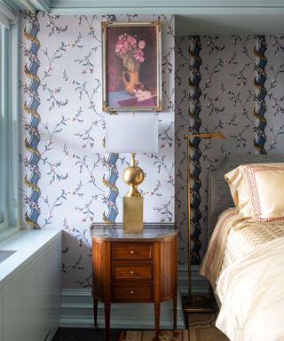 A blue bedroom with blue climbing floral and columnal wallpaper, a vintage wooden nightstand with gold art deco lamp, and a cream bed with silky patterned sheets
