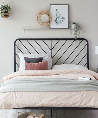 A bed with a black metal headboard, two gray pillows and a throw, a light pink sheet, with a white shelf with a mirror, framed wall art print, and a light green plant on it