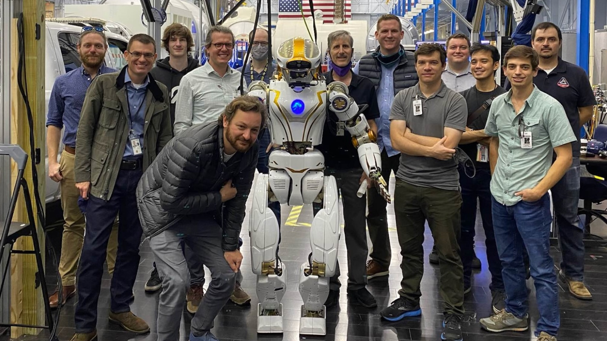 Twelve people stand near a humanoid robot inside a large hangar.