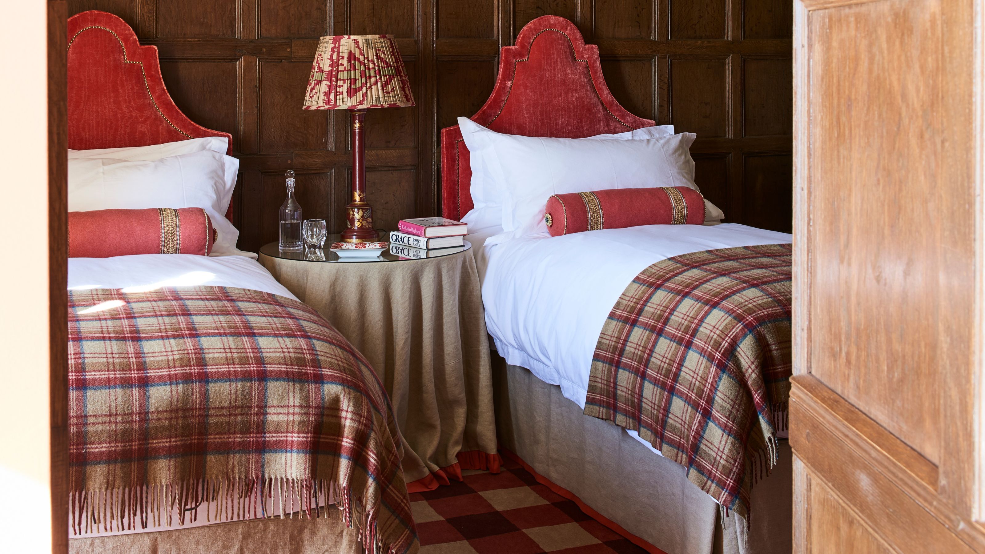 Wood panelled twin bedroom with single beds with red headboards, tartan blankets, and tartan carpet with a circular nightstand between the two beds with a patterned lamp and stack of books