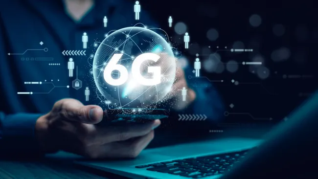 Una persona sosteniendo un teléfono móvil con la representación digital de un globo holográfico que contiene el texto "6G". En el fondo, se pueden ver íconos que simbolizan la conectividad y la interacción digital, como figuras de personas y elementos relacionados con redes y datos. El entorno es oscuro, destacando la pantalla y los elementos tecnológicos. La imagen refleja el concepto de la próxima generación de tecnología móvil, 6G, y su potencial para revolucionar la conectividad.