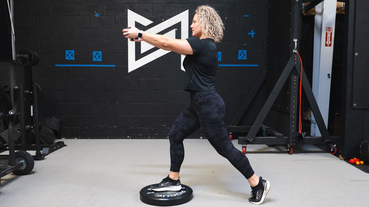 Woman stands in gym, with one foot on a weight plate on the floor and the other behind her, with her arms extended in front of her. She wears a black T-shirt, black leggings and black trainers.