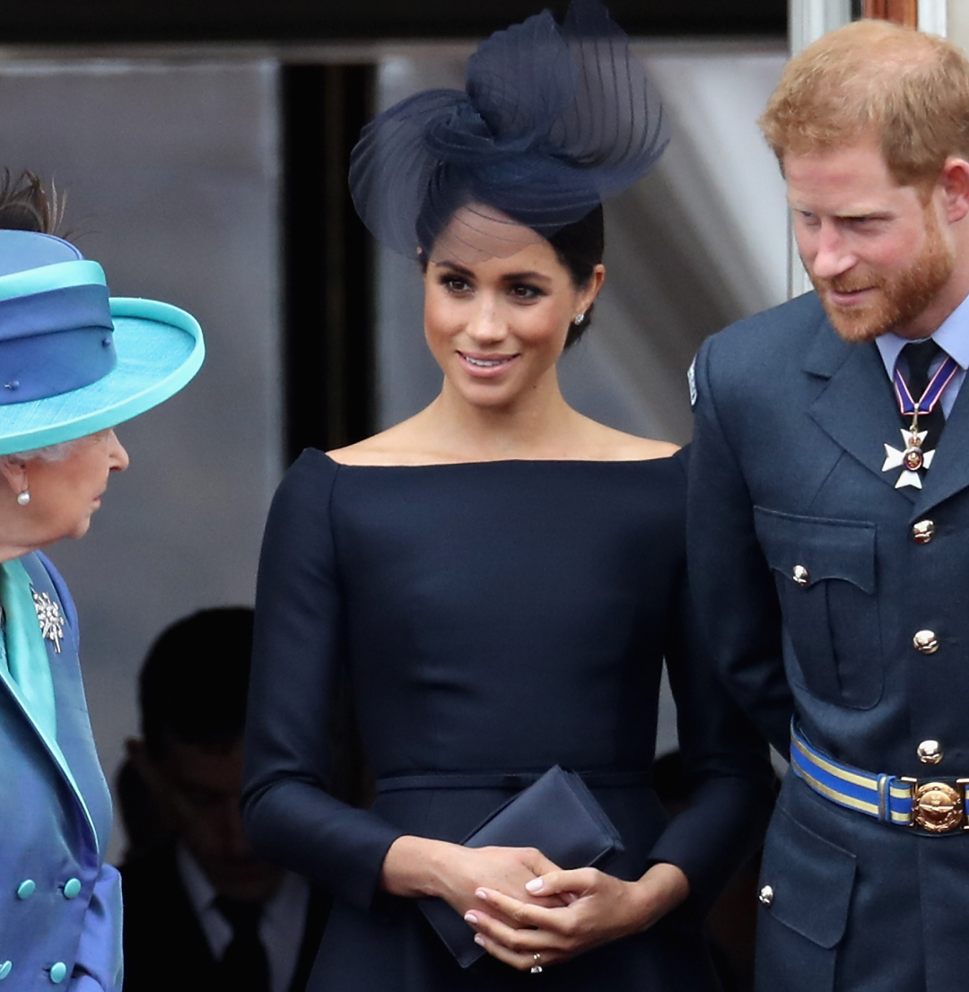 Queen Elizabeth wearing a blue coat standing next to Meghan Markle, in a navy dress and hat, and Prince Harry in a military uniform